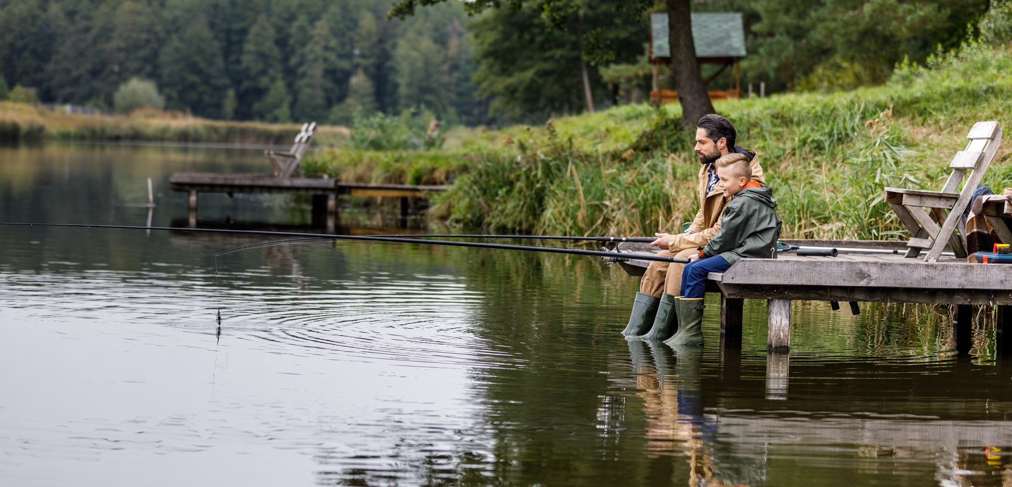 Vader en zoon zitten samen te vissen aan de rand van een visvijver in Limburg, omringd door groene natuur.