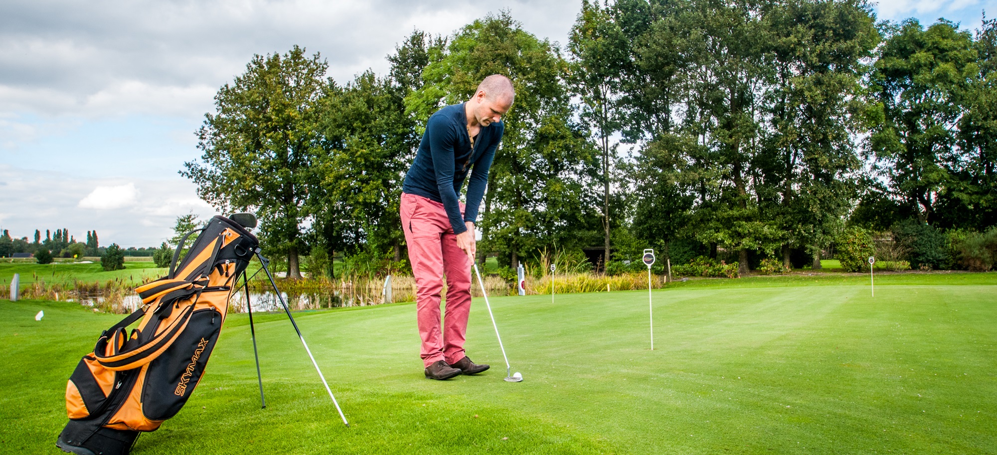 Man oefent zijn putt op een groene golfbaan met bomen op de achtergrond en een golftas op de voorgrond.