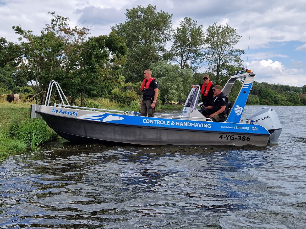 Controle- en handhavingsboot "De Remunj" van Sportvisserij Limburg vaart langs de oever van een waterplas. Drie handhavers in zwarte uniformen en met reddingsvesten staan aan boord. Op de achtergrond grazen runderen in een groen natuurgebied. De boot heeft een blauwe opdruk met de tekst "Controle & Handhaving" en het kenteken 4-YG-386.