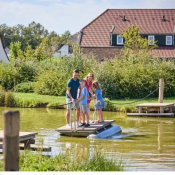 Gezin steekt met een vlot een vijver over op het parkterrein van de Leistert, met vakantiewoningen en groen op de achtergrond.