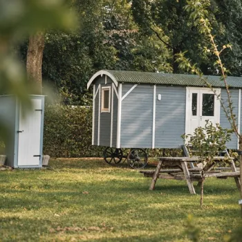 Grijsblauwe Shepherd Hut op wielen in Hunsel, met een houten picknicktafel op het gras en omringd door bomen en groen.
